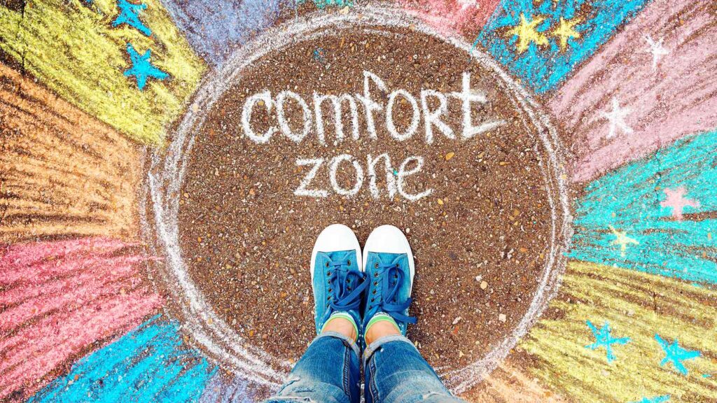 Image of a person with their feet inside of a circle sand with the words "comfort zone" included inside the circle.