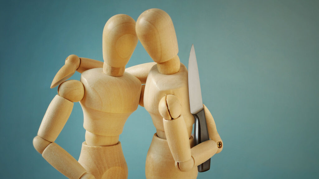 Two wooden artist mannequins embracing, with one holding a knife behind the other's back, set against a teal background. The embrace represents the appearance of closeness and trust, while the hidden knife represents the subtle betrayals happening beneath the surface. The danger is invisible to the person being held, mirroring how micro-betrayals operate undetected within relationships that still look intact.