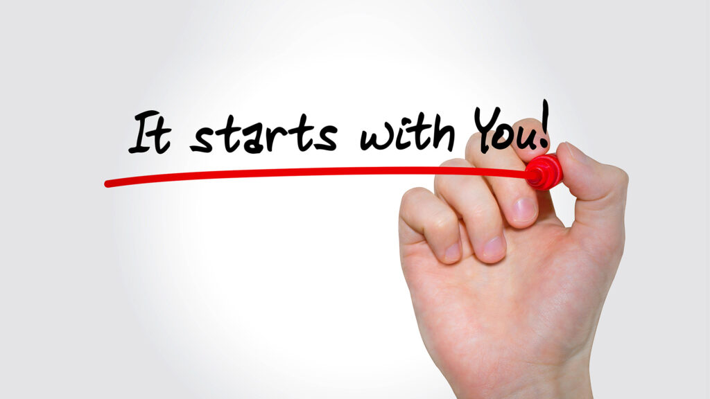 A hand holding a red marker writes the phrase "It starts with You!" on a transparent surface, with a bold red underline beneath the text on a clean white background. The image illustrates the concept of personal accountability and self-initiated growth, emphasizing that meaningful change and development begin with individual ownership rather than external circumstances.