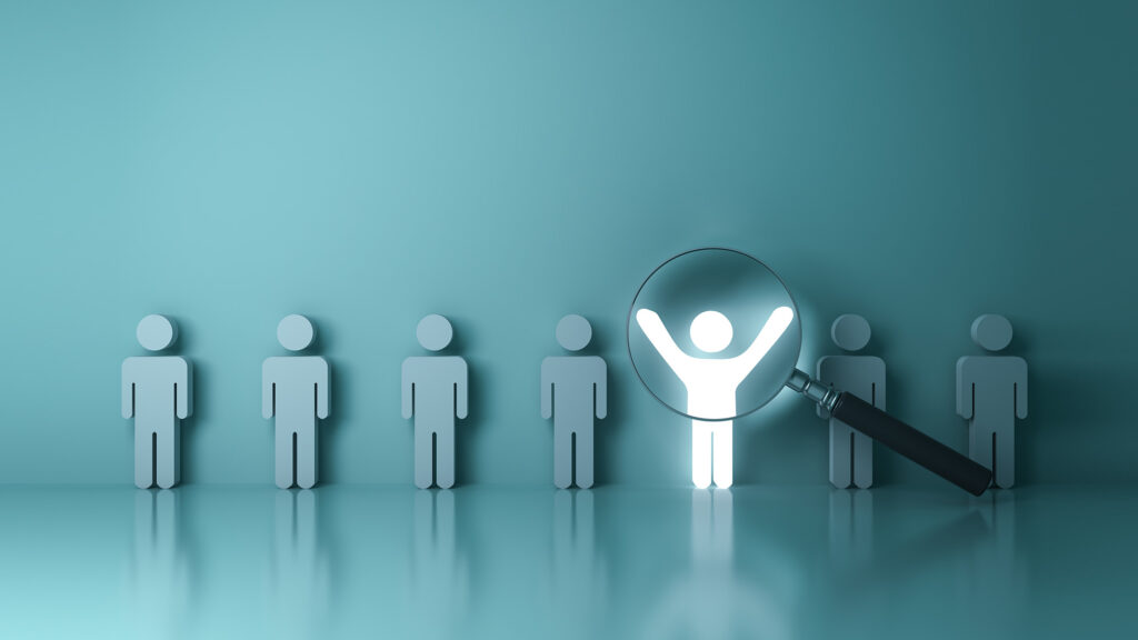 A row of identical grey human figures stands in a line against a teal background, with one glowing white figure raising its arms in celebration while highlighted by a magnifying glass. The image illustrates the concept of identifying and elevating standout talent in leadership, emphasizing the value of recognizing distinctive contributors who shine beyond the uniformity of the crowd.