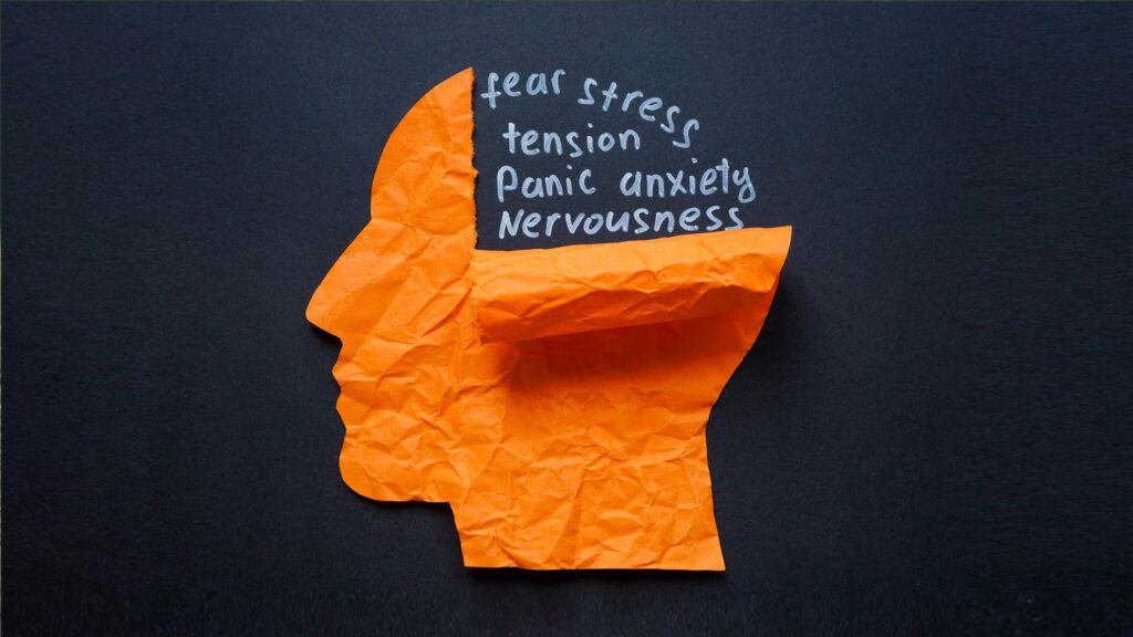 A crumpled orange paper cutout shaped like a human head in profile rests against a dark charcoal background, with the top of the head torn open to expose the dark space inside. Written in white chalk style handwriting within the opening are the words "fear, stress, tension, panic, anxiety, nervousness." The concept represents the internal weight of unprocessed emotions, illustrating how anxiety and stress crowd the mind when there is no space to slow down and respond rather than react.