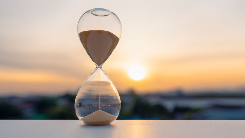A glass hourglass sits on a pale surface, sand falling steadily from the top chamber into the bottom. Behind it, the sun sets low over a blurred city skyline in warm golden light. The image represents the passage of time and the weight of decisions made within it. The setting sun suggests that time is finite and that how it is used carries lasting consequence.