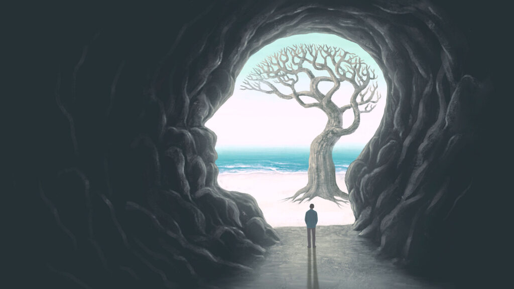 A lone figure stands at the mouth of a dark cave that is shaped like a human head in profile. Beyond the opening, a solitary tree stands against a soft pale sky, with the ocean and a stretch of beach behind it. The image represents inner exploration and personal growth, with the cave as the inner mind and the tree beyond it as potential waiting to be reached. The small figure suggests the journey from internal reflection toward open possibility.