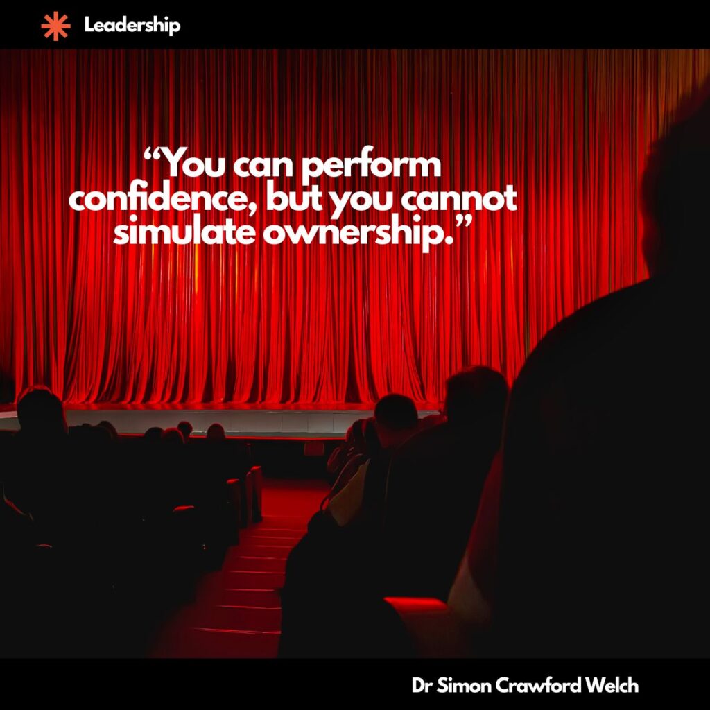 The silhouetted heads and shoulders of an audience are visible in the foreground of a darkened theater, looking toward a brightly lit stage framed by tall, rich red velvet curtains that dominate the scene. The image illustrates the concept of performance versus authenticity in leadership, contrasting the staged presentation of confidence with the unseen reality of genuine accountability. Quote over the image reads, "You can perform confidence, but you cannot simulate ownership." By Dr. Simon Crawford Welch.