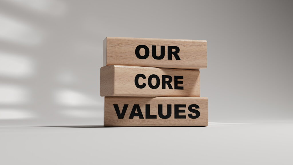 Visual description: Three light wood blocks stacked slightly askew against a plain white studio backdrop, printed in bold black capitals with the words OUR, CORE, and VALUES, with soft window light casting a diagonal shadow behind them. Concept: Values stated aloud are easy to stack up and display, but the blocks sit unevenly, hinting that what we claim to stand for and how we actually behave rarely line up without honest self examination.