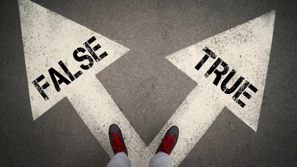 Visual description: An overhead view of a person standing on grey asphalt in brown boots with red laces, feet planted at the point where two large painted white arrows diverge, the left arrow stencilled FALSE and the right arrow stencilled TRUE. Concept: Every leader stands at this junction more often than they admit, and the direction chosen in small unwitnessed moments is what a culture eventually learns to imitate.