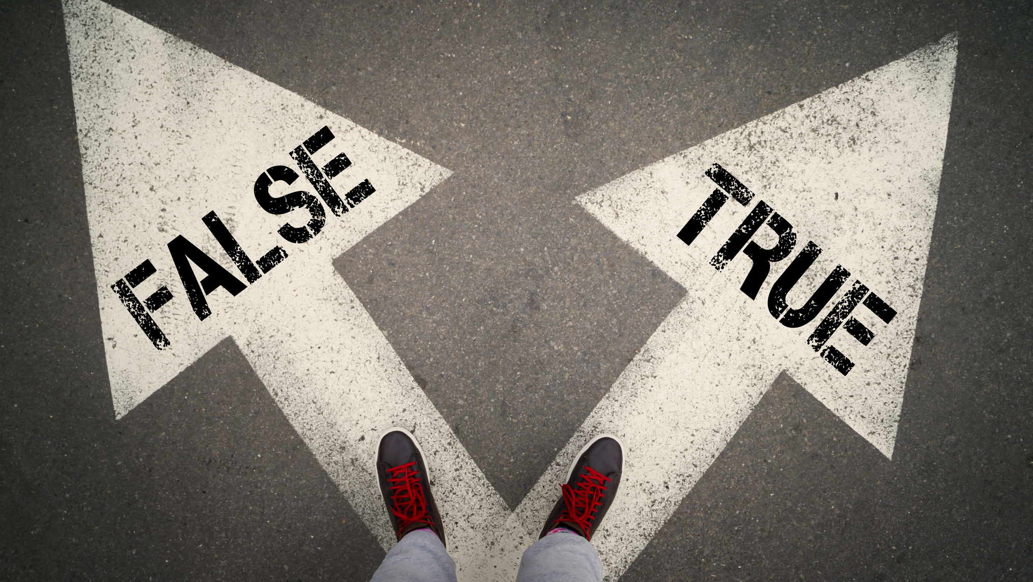 Visual description: An overhead view of a person standing on grey asphalt in brown boots with red laces, feet planted at the point where two large painted white arrows diverge, the left arrow stencilled FALSE and the right arrow stencilled TRUE. Concept: Every leader stands at this junction more often than they admit, and the direction chosen in small unwitnessed moments is what a culture eventually learns to imitate.