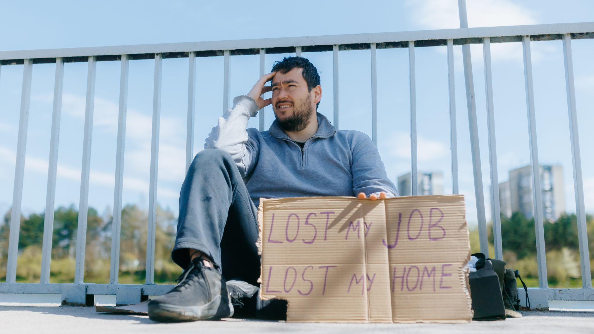Visual description: A bearded man in a grey sweatshirt sits on concrete against a white railing under a bright blue sky, one hand pressed to his forehead, a hand lettered cardboard sign propped in front of him reading LOST MY JOB LOST MY HOME, with a backpack beside him and city towers in the distance. Concept: The hardest chapters arrive without warning and without dignity, and they look nothing like the polished growth stories we are sold. Development happens inside the wreckage, not after it has been cleaned up.