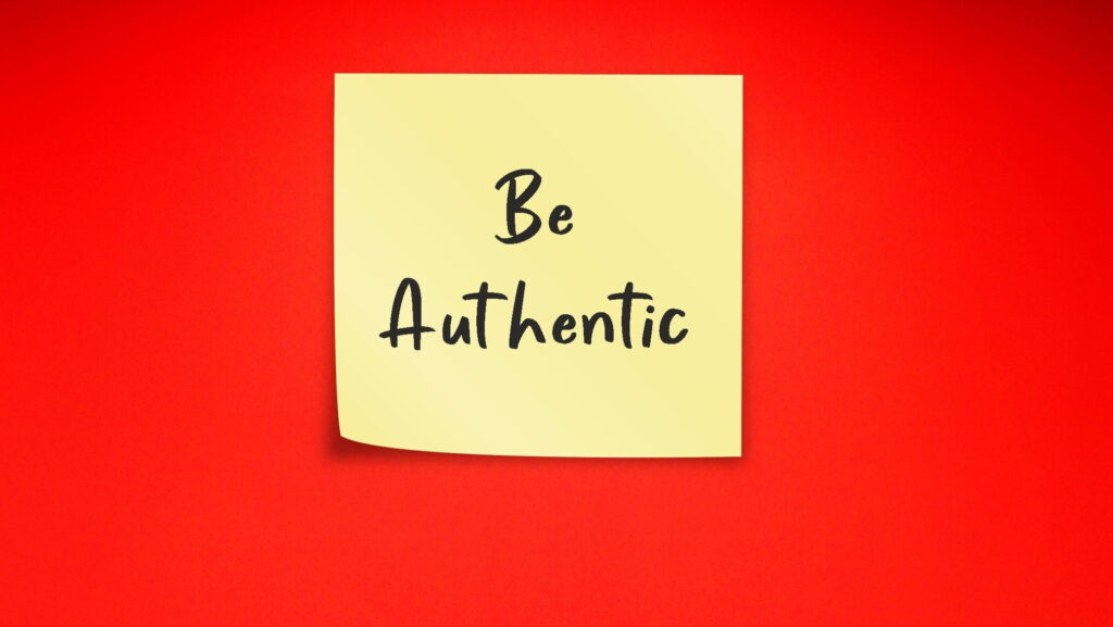 Visual description: A pale yellow sticky note attached to a bright red wall, curling slightly at the lower left corner, with the handwritten words "Be Authentic" in black marker. Concept: The reminder note is the smallest possible instruction and the hardest one to follow, suggesting that authenticity is less a personality trait than a daily practice we have to keep posting back to ourselves.