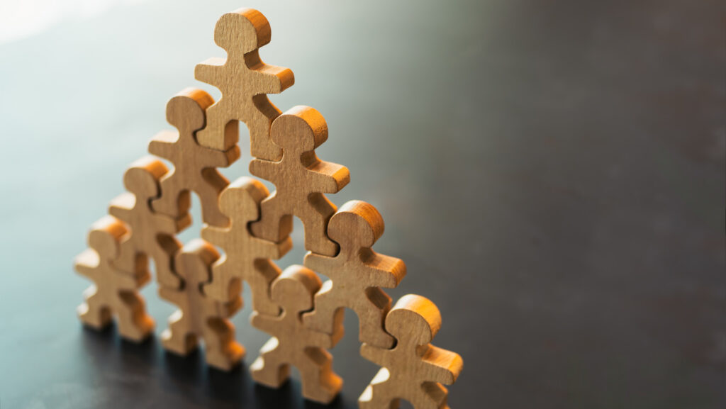 Wooden puzzle figures shaped like people interlocked and stacked into a pyramid on a dark surface, lit warmly from above with soft shadows behind them. The image represents structure built from connection, where leadership holds only because each piece is genuinely fitted to the ones beside and beneath it.