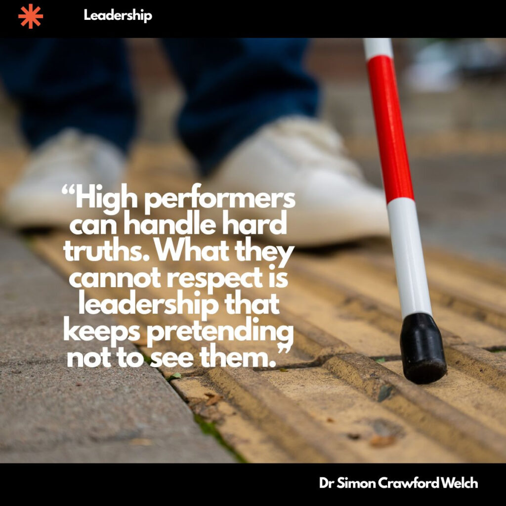 A red and white cane tip rests on a yellow tactile paving strip on a sidewalk, with a person's feet in white shoes and blue jeans blurred in the background. The concept is noticing what is really in front of you and refusing to look past it. Text over the image reads: "High performers can handle hard truths. What they cannot respect is leadership that keeps pretending not to see them." By Dr Simon Crawford Welch.
