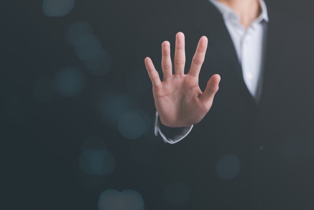 A person in a dark suit and white shirt, face out of frame, holds an open palm up toward the camera in a stop gesture against a dark blurred background with soft bokeh lights. The image represents the reflexive hand a defensive leader raises against feedback, and what that gesture teaches everyone watching.