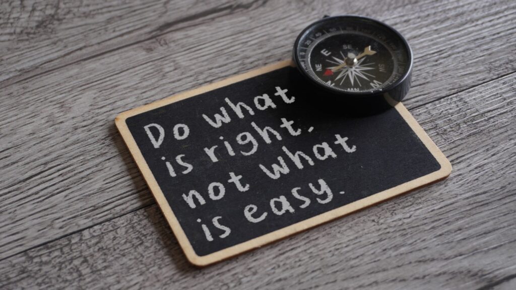 A small black chalkboard lies on a grey weathered wood surface with the words Do what is right, not what is easy handwritten in white chalk, and a black pocket compass rests on its top corner. The image represents the discipline of choosing truth over comfort as power makes the easy story more tempting.