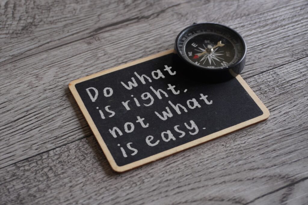 A small black chalkboard lies on a grey weathered wood surface with the words Do what is right, not what is easy handwritten in white chalk, and a black pocket compass rests on its top corner. The image represents the discipline of choosing truth over comfort as power makes the easy story more tempting.