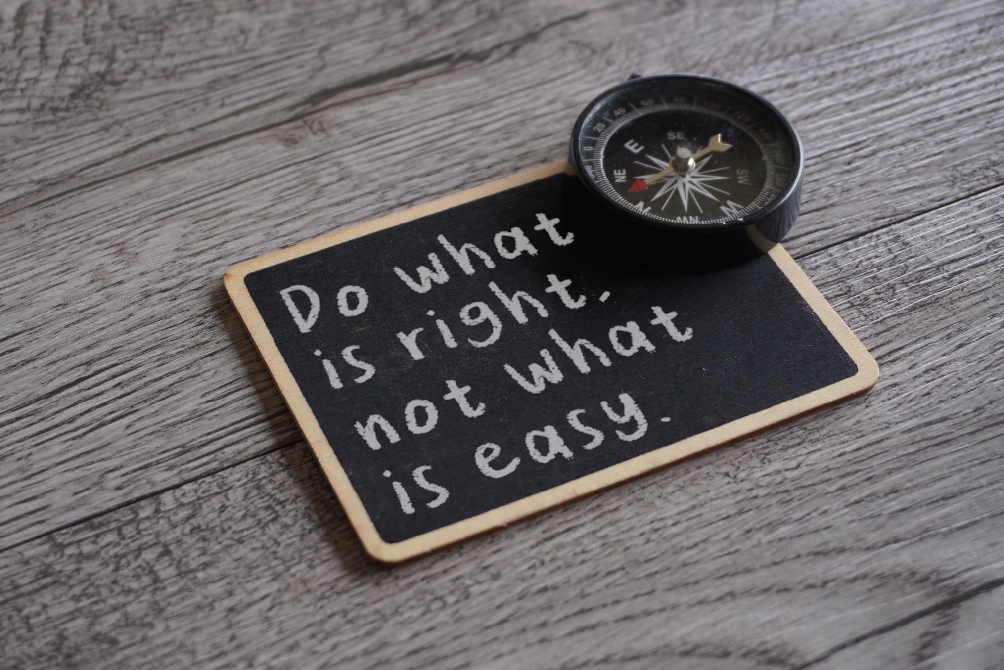 A small black chalkboard lies on a grey weathered wood surface with the words Do what is right, not what is easy handwritten in white chalk, and a black pocket compass rests on its top corner. The image represents the discipline of choosing truth over comfort as power makes the easy story more tempting.