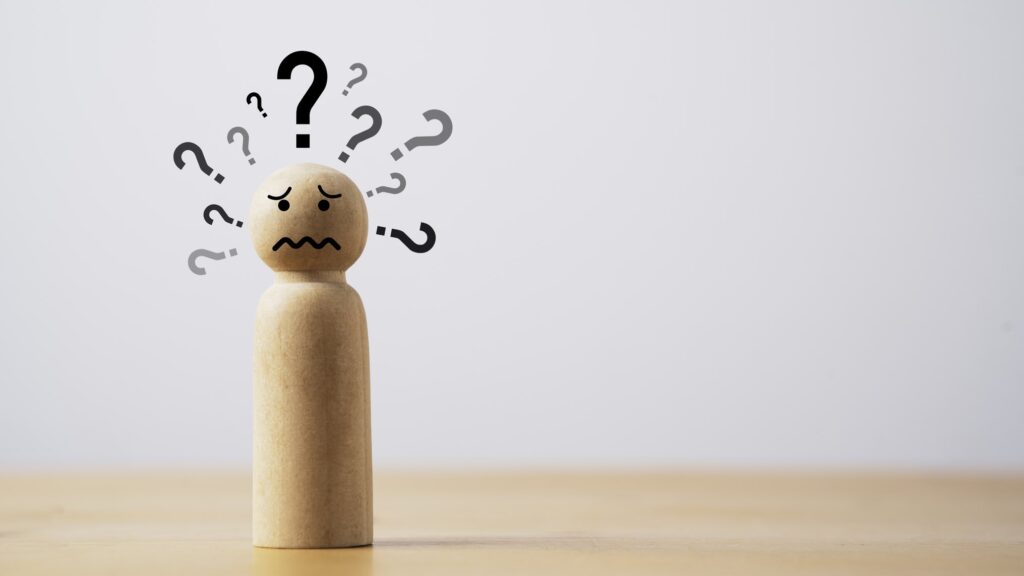A small wooden peg figure with a worried face drawn on its head stands on a light wooden table against a plain grey wall, surrounded by black and grey question marks. The image represents the uncertainty that forceful people rush to silence, and why volume so often gets mistaken for authority.
