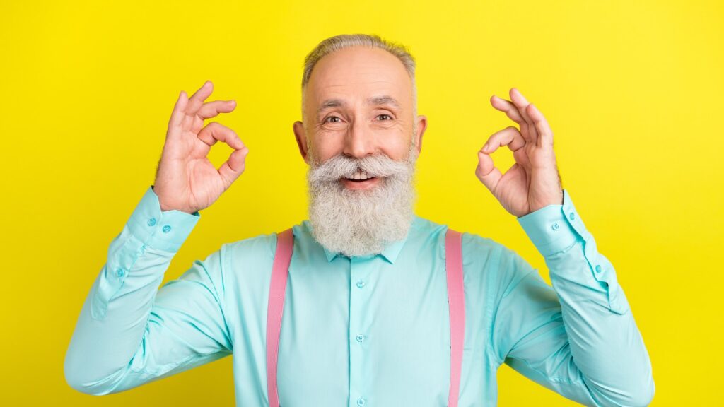 A smiling older man with a full white beard, a mint green shirt and pink suspenders holds up both hands in an OK sign against a bright yellow background. The image represents the polished, everything is fine confidence that can start to feel less believable than an honest admission of uncertainty.