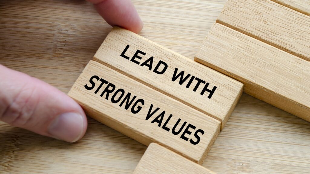 A hand slides two light wooden blocks into place on a bamboo surface, printed in bold black letters with the words Lead With Strong Values, beside a row of plain blocks. The image represents leadership that is built on what is true rather than on the story an organization prefers to tell.