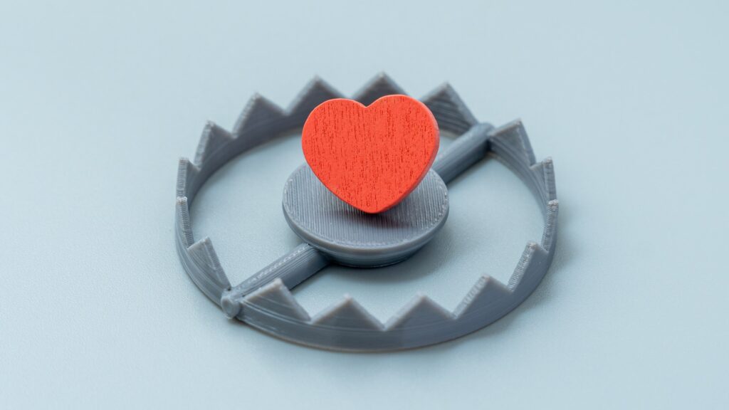 A red wooden heart sits on the trigger plate of a small grey toothed trap against a pale blue grey background. The image represents how a toxic relationship can hold your heart in place long after it has started to cause harm.