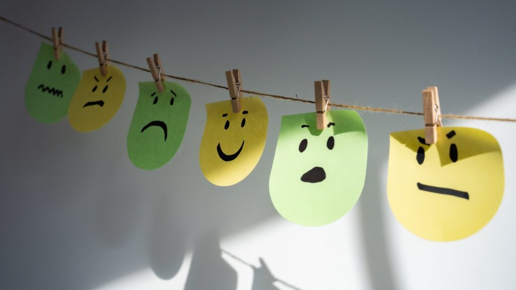 Six paper faces in green and yellow hang from a twine line by wooden clothespins against a pale grey wall, each drawn with a different expression, from anxious and angry to sad, happy, surprised and annoyed, with their shadows falling on the wall below. The image represents the range of emotions we feel and the need to name each one precisely.