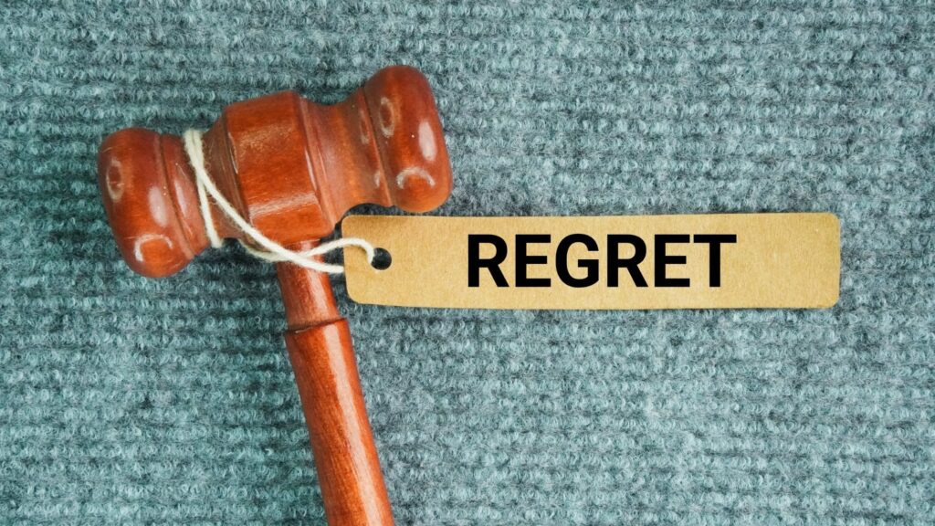 A polished wooden gavel lies on a teal grey carpet with a kraft paper tag tied to its handle that reads REGRET in bold black letters. The image represents regret as a verdict we pass on ourselves, and the choice to learn from it rather than repeat it.
