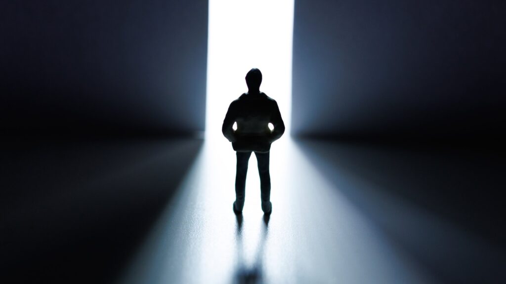 A small figurine of a man in a hooded jacket stands in silhouette, hands in his pockets, before a tall, narrow opening of bright white light between two dark walls, his shadow stretching toward the viewer. The image represents stepping toward a new self while the old story still trails behind.
