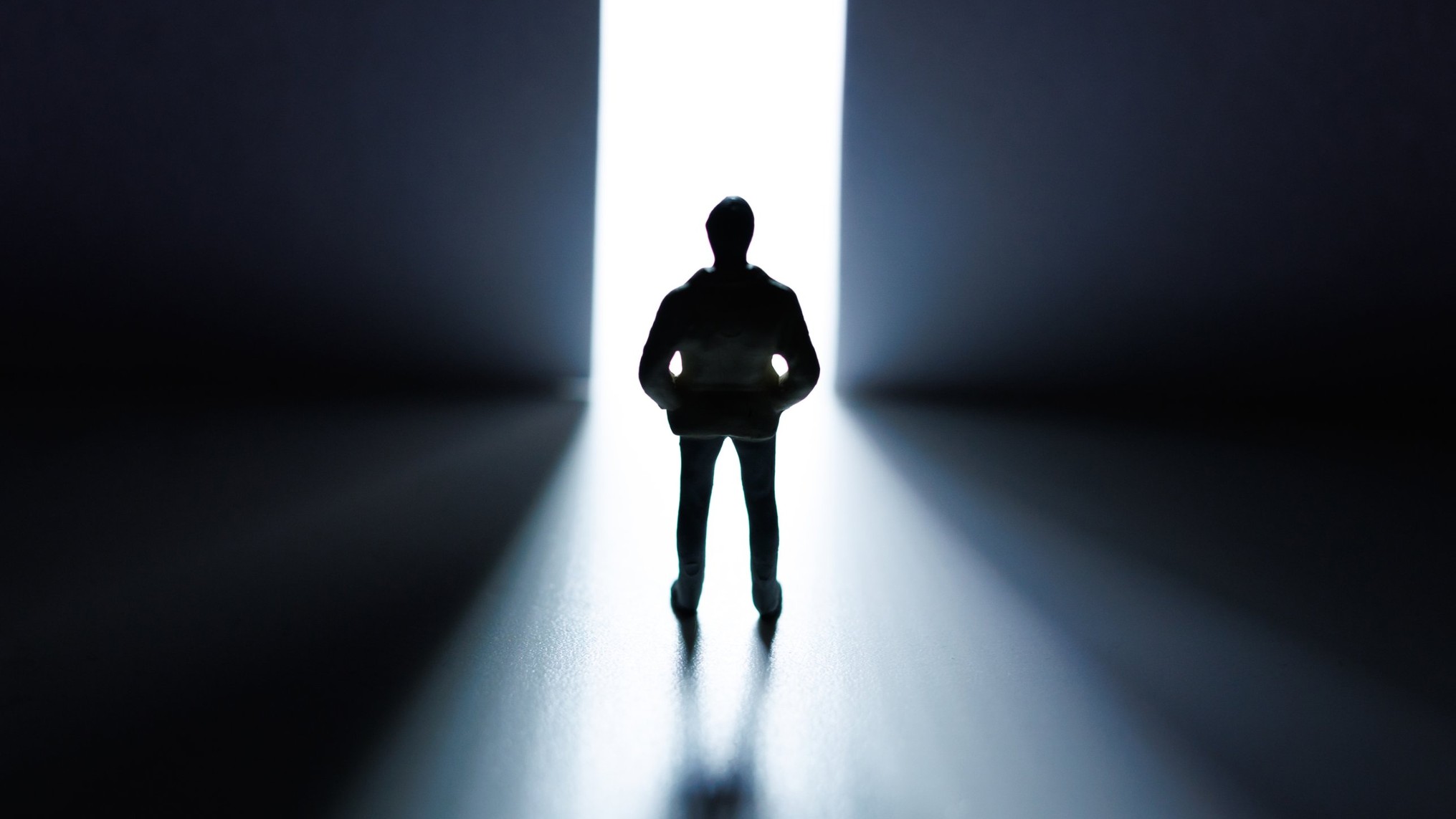 A small figurine of a man in a hooded jacket stands in silhouette, hands in his pockets, before a tall, narrow opening of bright white light between two dark walls, his shadow stretching toward the viewer. The image represents stepping toward a new self while the old story still trails behind.