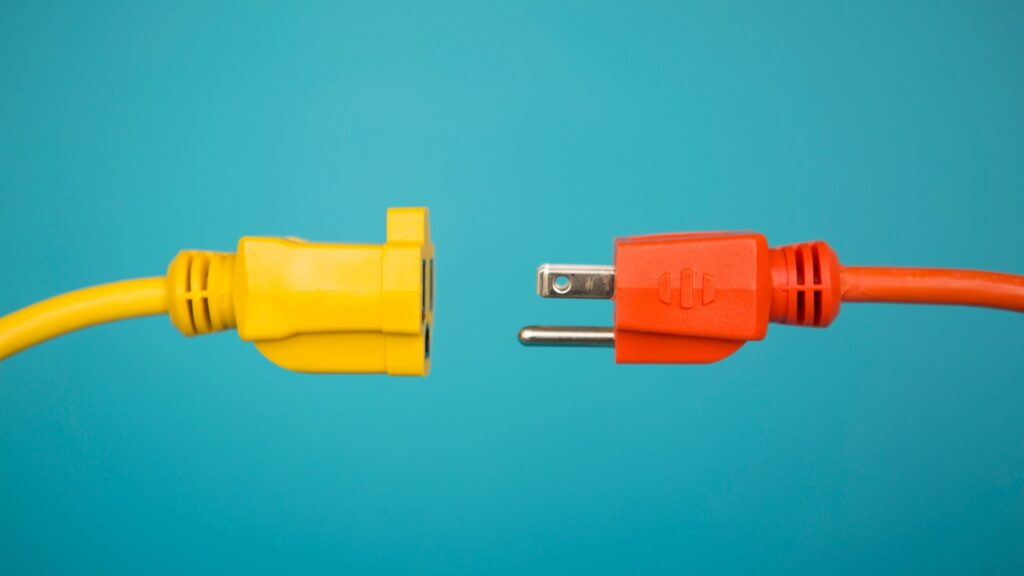 A yellow extension cord socket and an orange plug face each other against a teal background, almost touching but not connected. The image represents the gap between refining the system and doing the work it was built for.