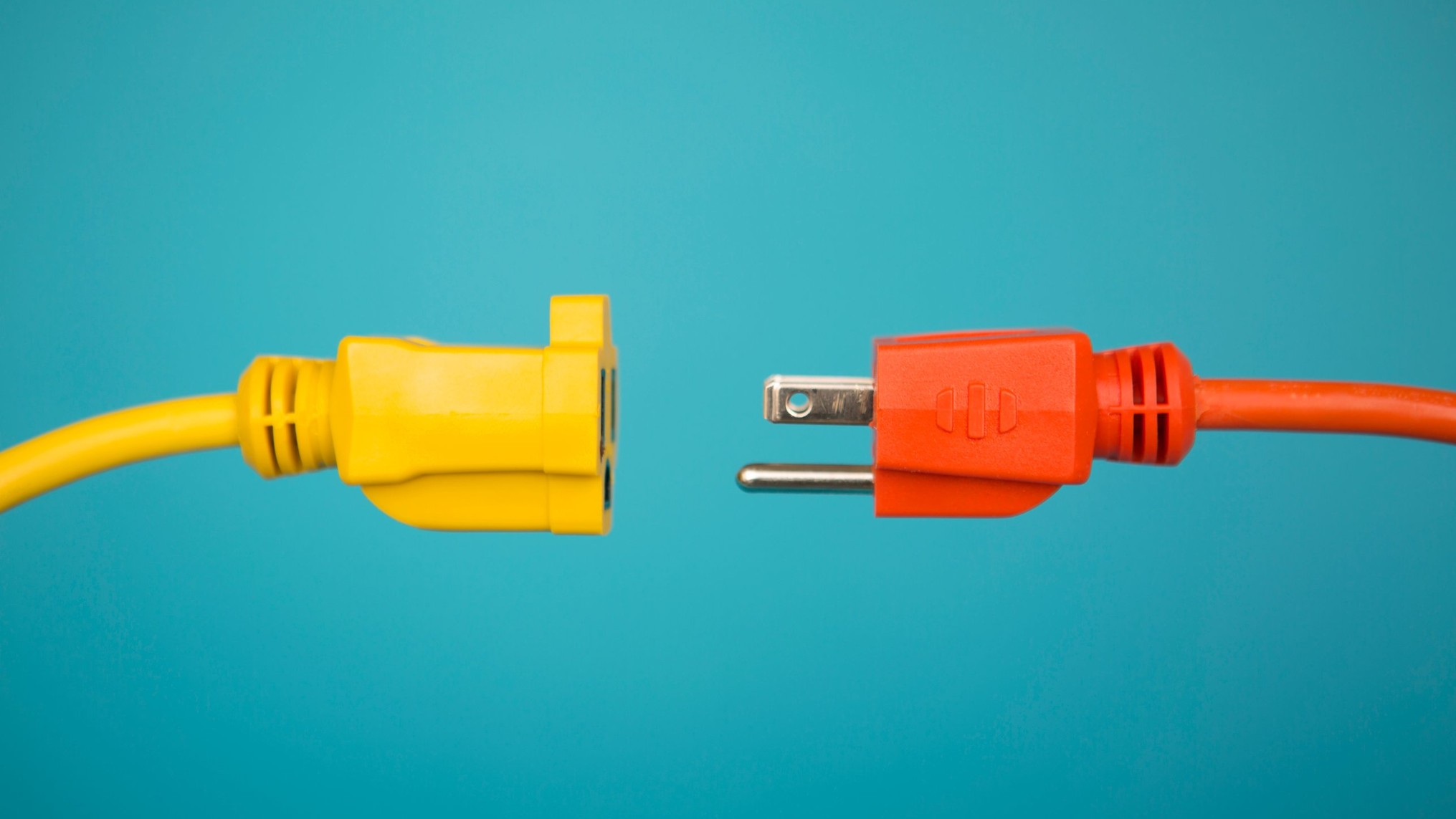 A yellow extension cord socket and an orange plug face each other against a teal background, almost touching but not connected. The image represents the gap between refining the system and doing the work it was built for.