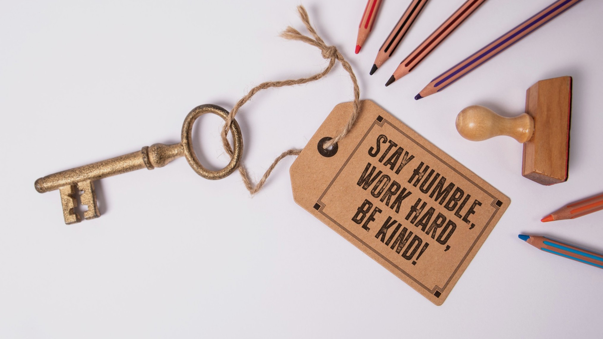 An antique brass key tied with twine to a brown paper tag stamped STAY HUMBLE, WORK HARD, BE KIND! rests on a light grey surface beside colored pencils and a wooden rubber stamp. The image represents humility as the key that keeps growth open.