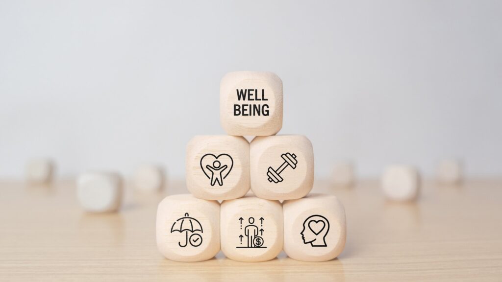 Six light wooden cubes are stacked in a pyramid on a pale wooden table against a soft grey wall, the top cube printed WELL BEING and the others marked with line icons of a person inside a heart, a dumbbell, an umbrella with a check mark, a figure with rising arrows and a coin, and a head with a heart inside. The image represents the inner world each of us is responsible for building and tending.