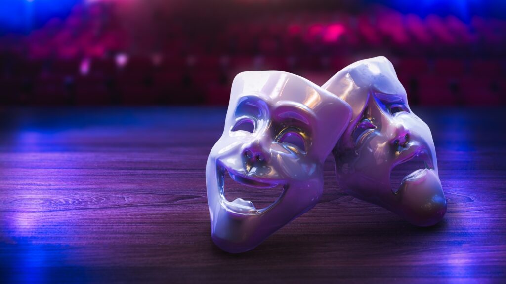 Two glossy white theater masks, one laughing and one weeping, rest on a wooden stage under purple and blue light, with the blurred red seats of an auditorium behind them. The image represents leadership that performs a role rather than living it.